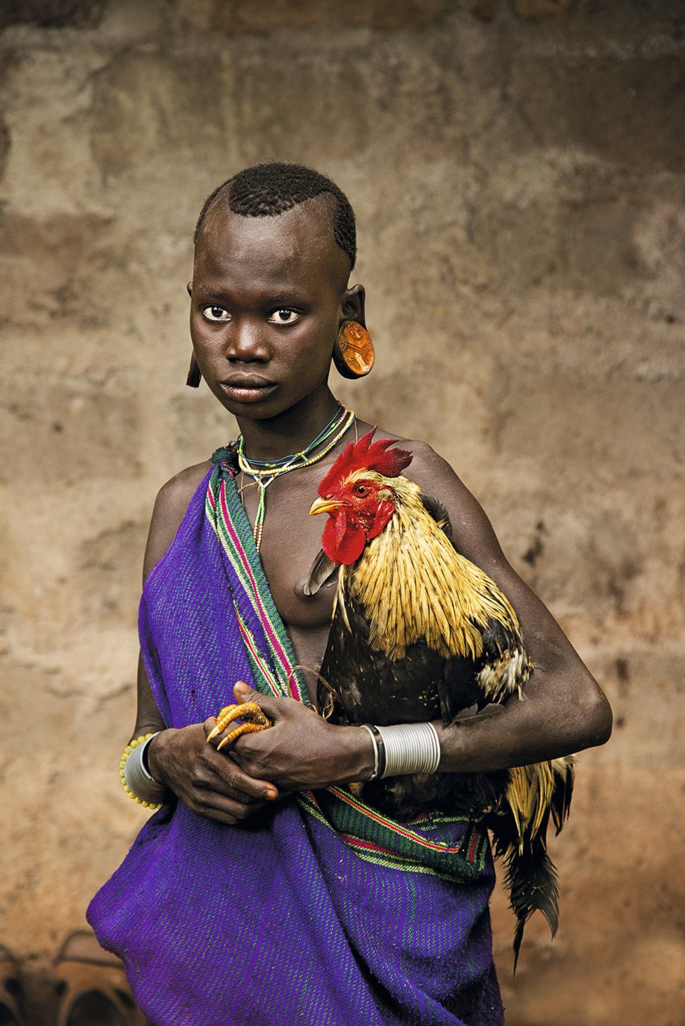 Una niña de la tribu kara del valle del Omo (Etiopía) sostiene un gallo. Las garras del animal asoman, pacíficas, entre las manos de la pequeña. Año 2013.
